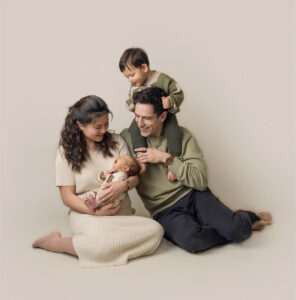Family of four sitting on almond-colored backdrop with mom holding newborn while toddler in on dad's shoulders. Mom wearing a cream outfit and dad and son are wearing an olive green colored shirt. 