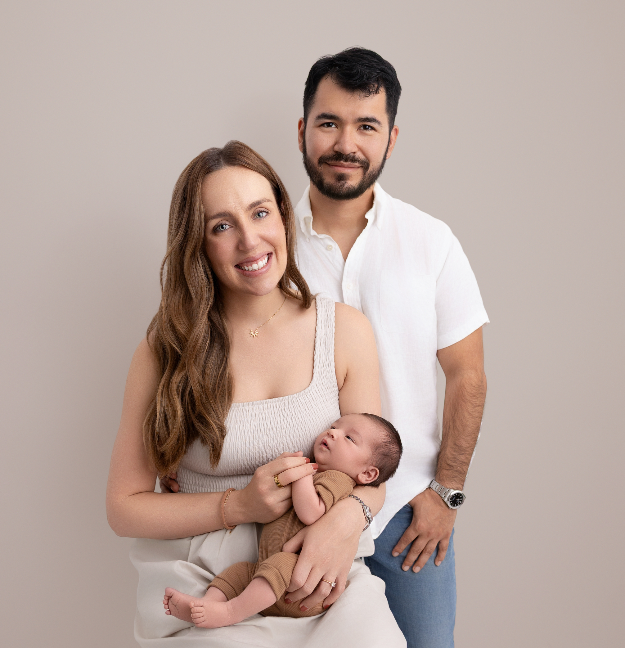 Family of three with mom wearing a khaki colored dress and holding newborn boy in her arms while sitting on a stool and dad is next to mom in a white shirt.