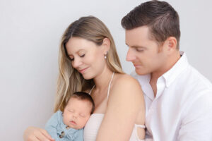 First time parents sitting posing against an off-white backdrop with their newborn baby wearing neutral colors and baby wearing a light blue outfit. 