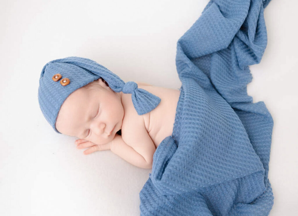 Newborn baby boy asleep and posed gently against a white blanket and wearing a baby blue hat and wrap posed for photo session.