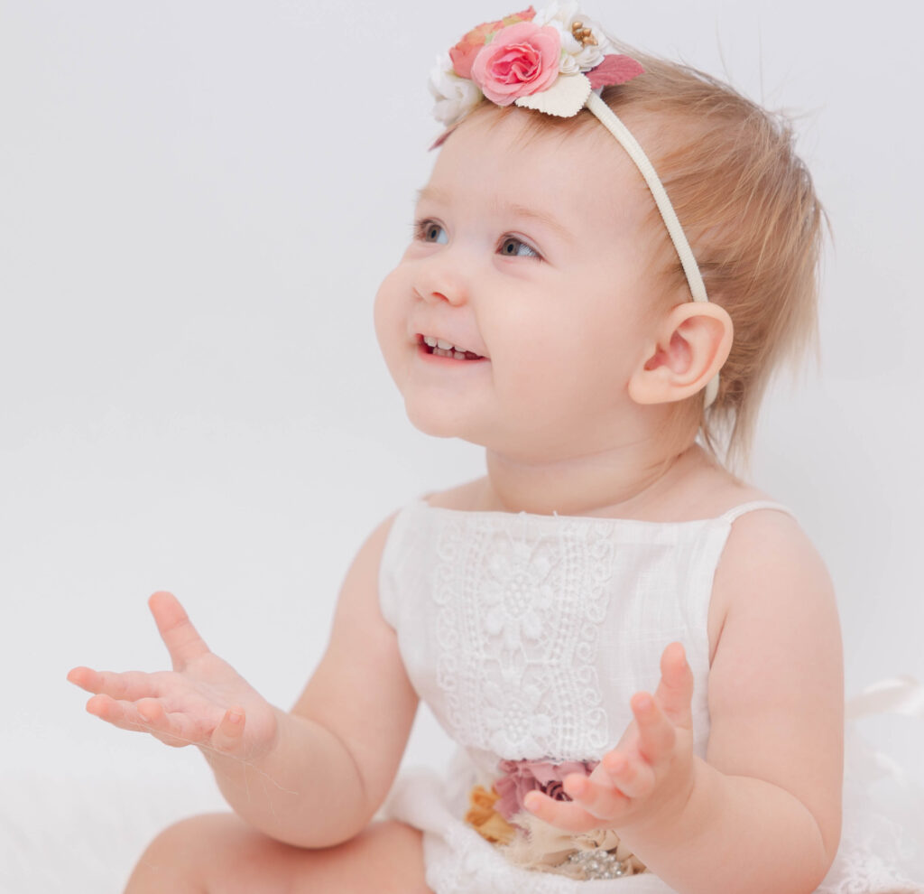 One-year-old girl wearing a white dress and flowered headband for her birthday photo session at Heather Nicole Photography.