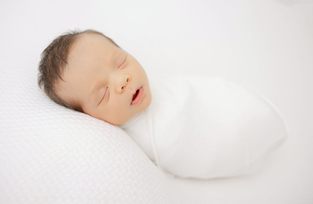 Newborn baby boy wrapped and posed on a white backdrop and sleeping soundly for photo session at Heather Nicole Photography Studio.