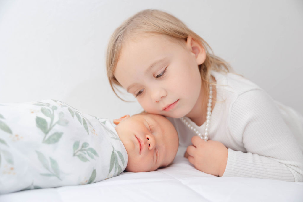 A toddler girl in a white shirt rests her cheek on her sleeping baby sibling's head after visiting baby boutiques in redondo beach