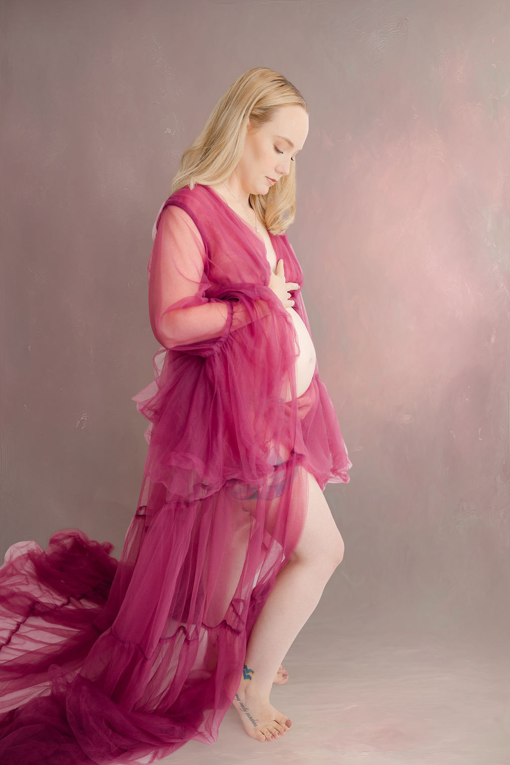 A pregnant woman stands in a studio with hands on her bump as she looks down to it in pink maternity gown after meeting doulas in redondo beach