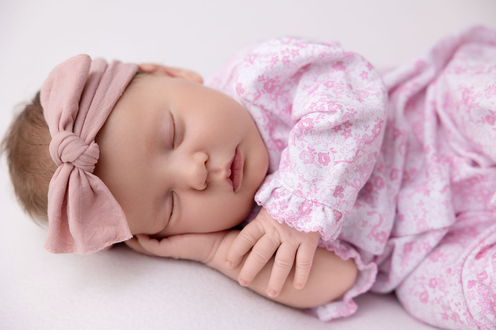 A newborn baby sleeps on her side in a pink flower print onesie and headband