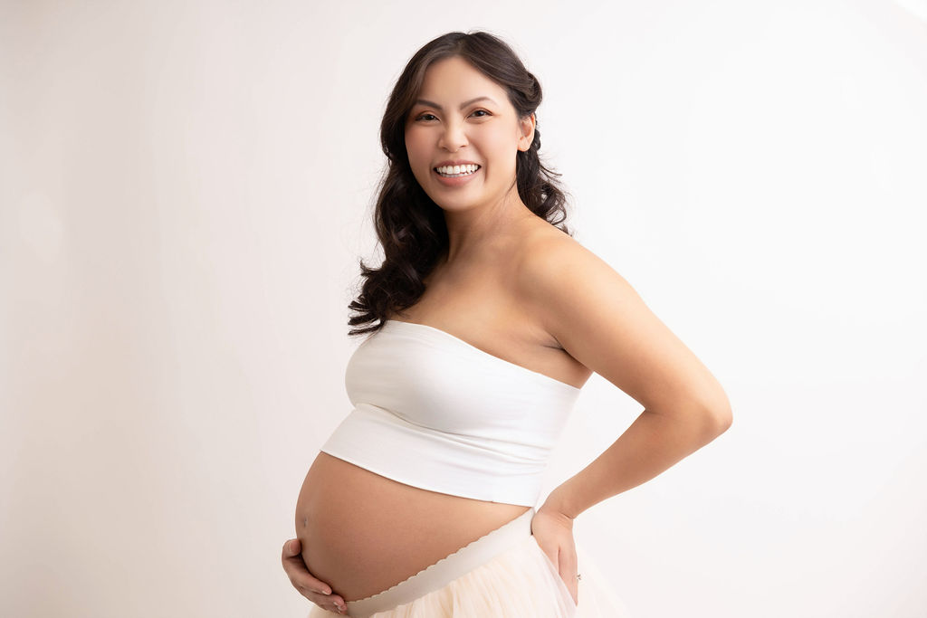 A happy pregnant woman smiles while standing in profile with a hand under her exposed bump