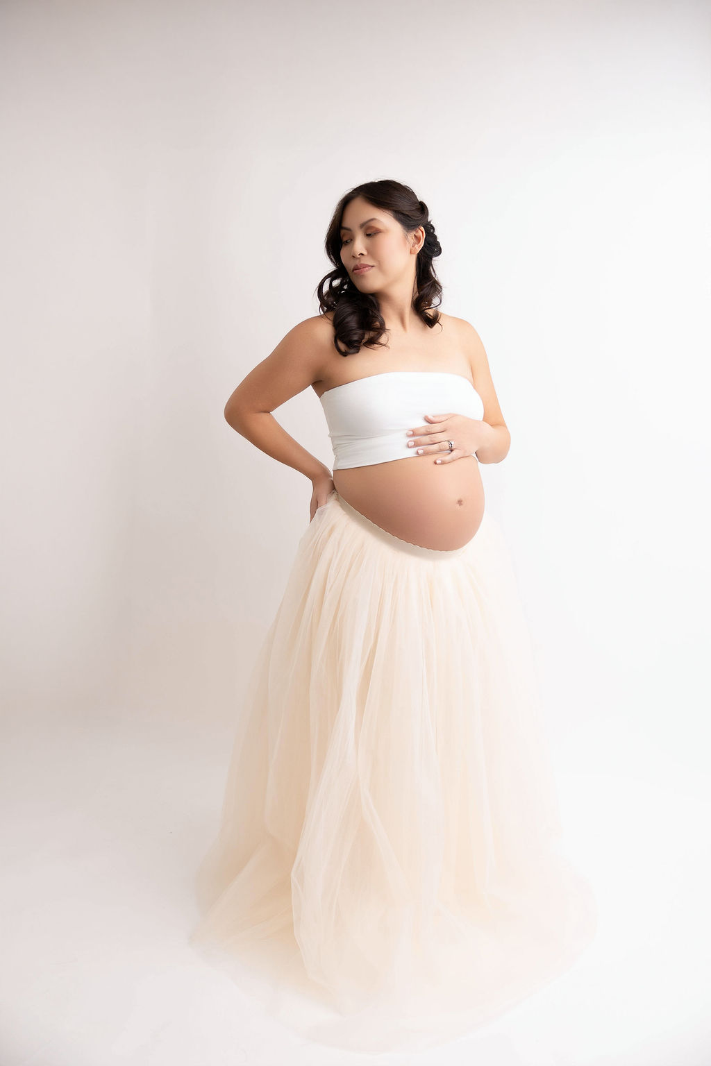 A mother to be stands in a studio with a hand on her bump and eyes closed in a white tube top and cream skirt after meeting midwives in redondo beach