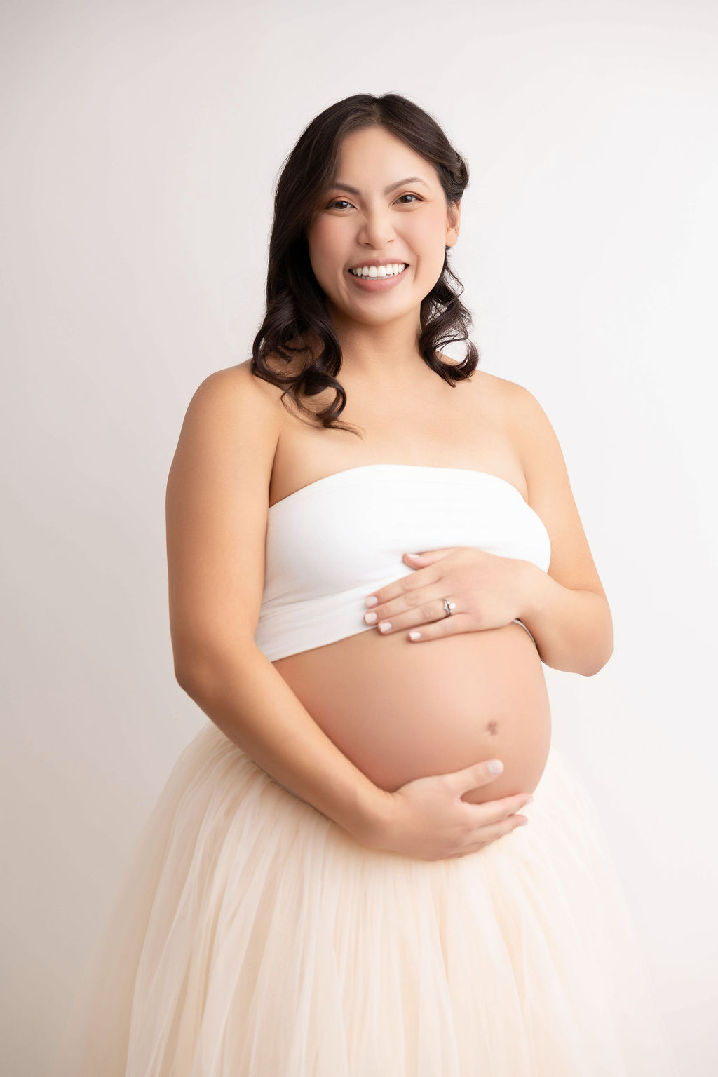A happy mother to be stands in a whtie tube top and cream skirt in a studio with hands on her bump after meeting midwives in redondo beach