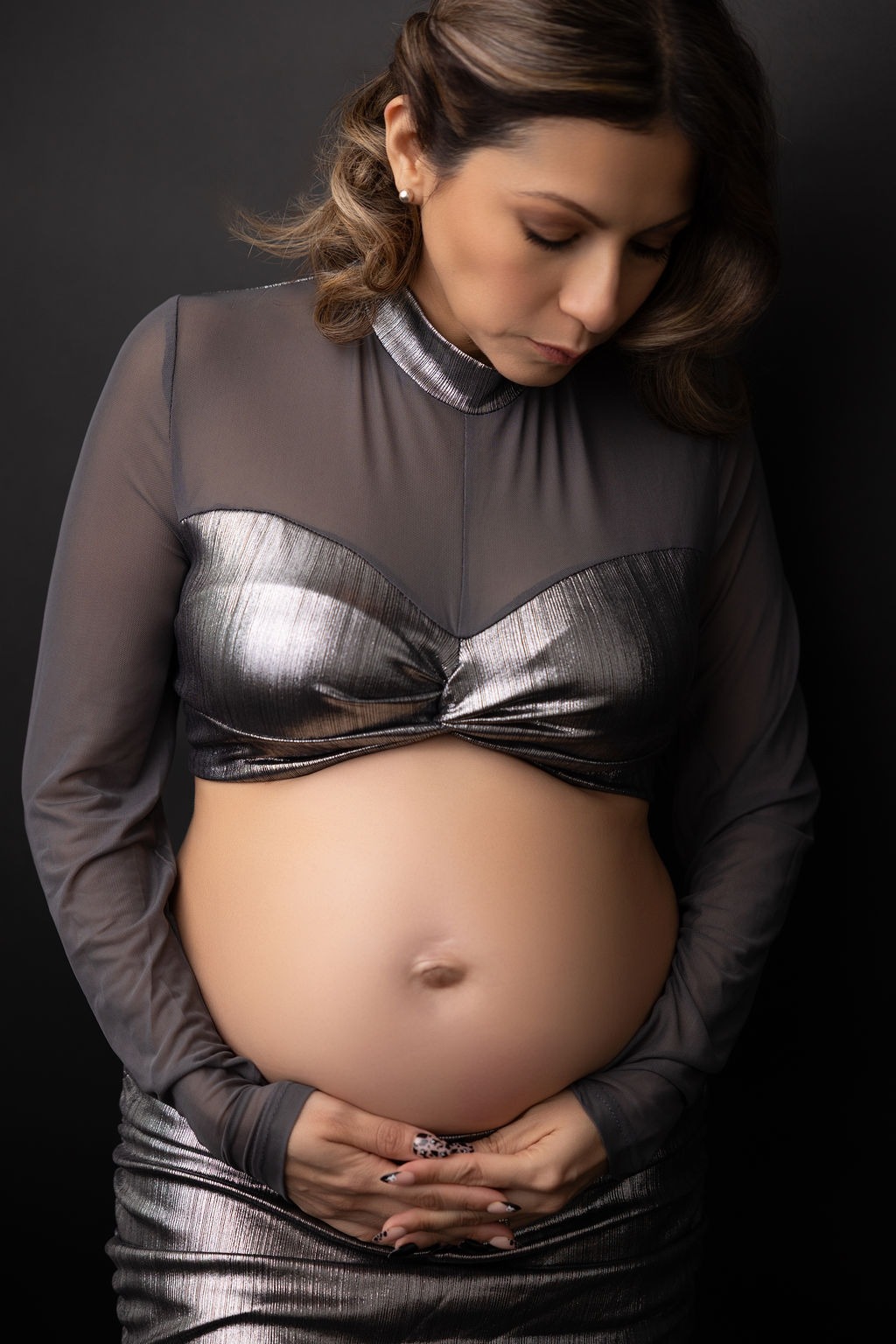 A mother to be looks down to her hands under her exposed bump in a silver gown