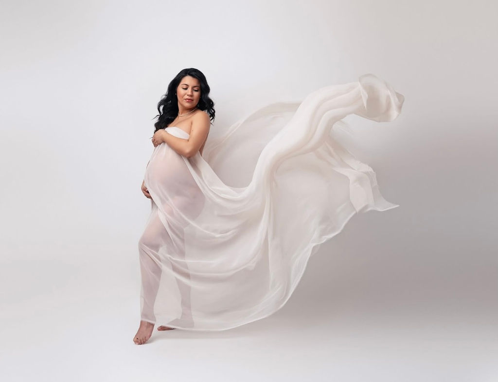 A happy pregnant woman stands in a studio wrapped ina. sheer white silk as it blows int he wind