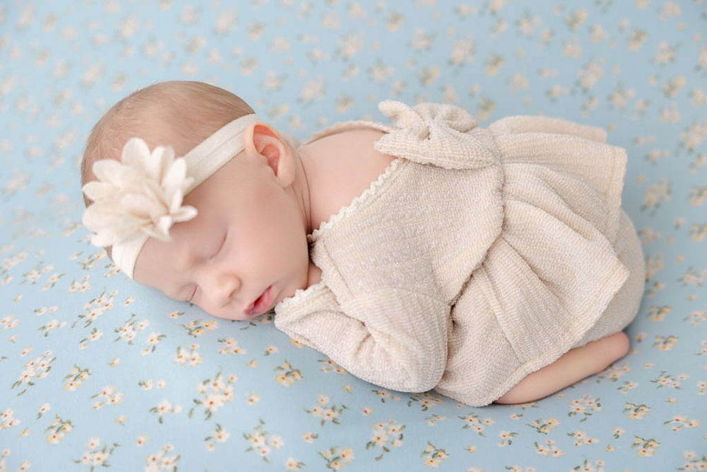 A newborn baby girl sleeps in froggy pose in a tan dress on a blue bed before visiting pediatric dentists in redondo beach