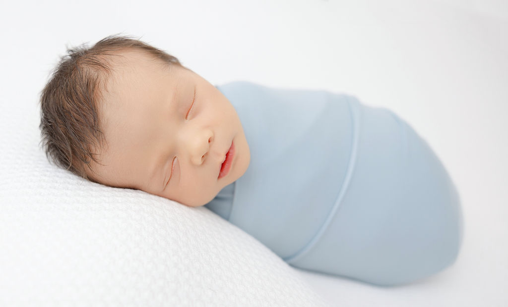 A newborn baby sleeps in a blue swaddle on a white bed after visiting pediatricians in redondo beach