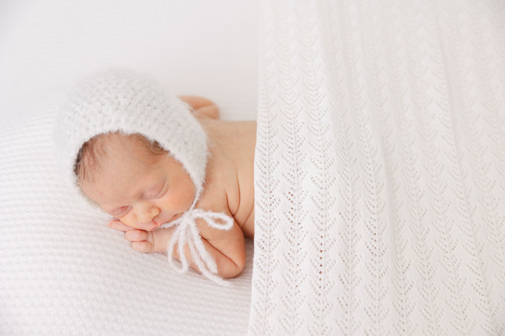 A newborn baby sleeps in a knit bonnet under a white blanket on its tummy after visiting daycares in redondo beach