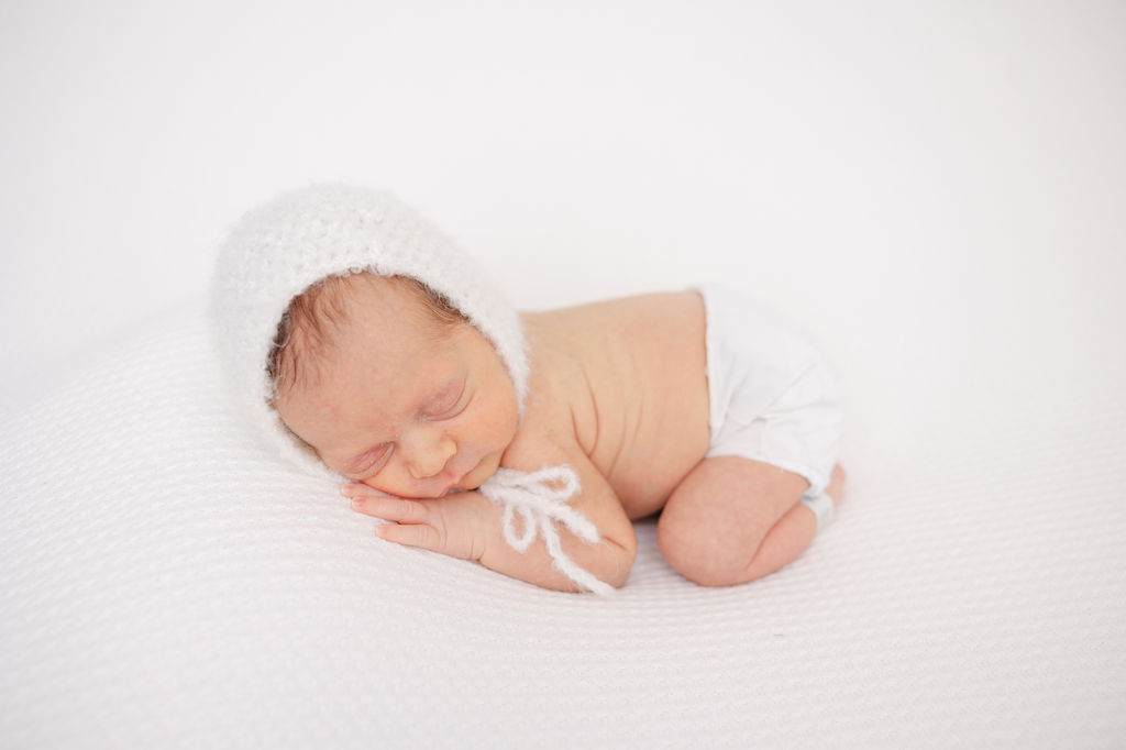 A sleeping newborn baby in a white bonnet in froggy pose on a white bed