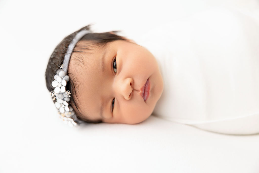 A closeup of a newborn baby laying in a white swaddle with one eye open before some infant swim lessons in redondo beach