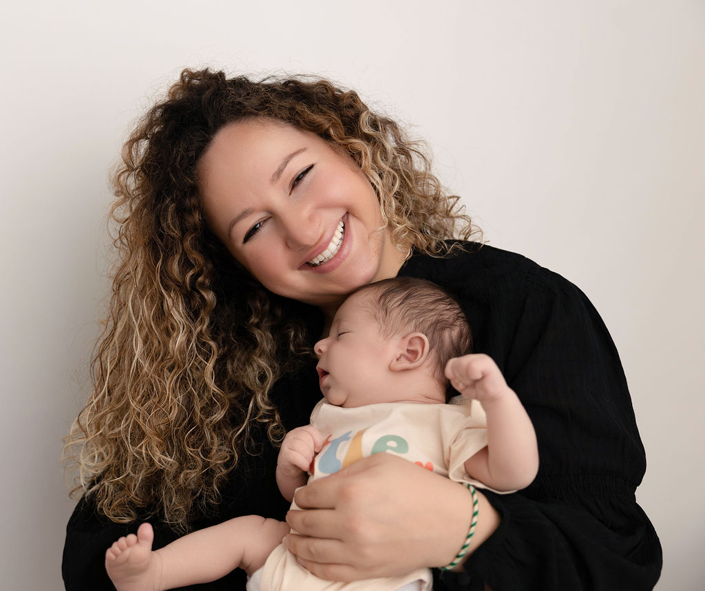 A smiling mother in a black sweater cradles her newborn sleeping against her chest in a studio