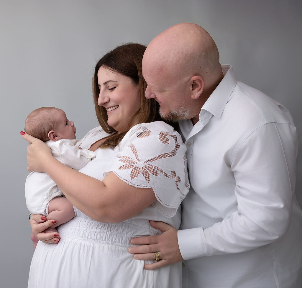 New mom and dad stand in white in a studio playing with their newborn baby against mom's chest after some mommy and me classes in redondo beach