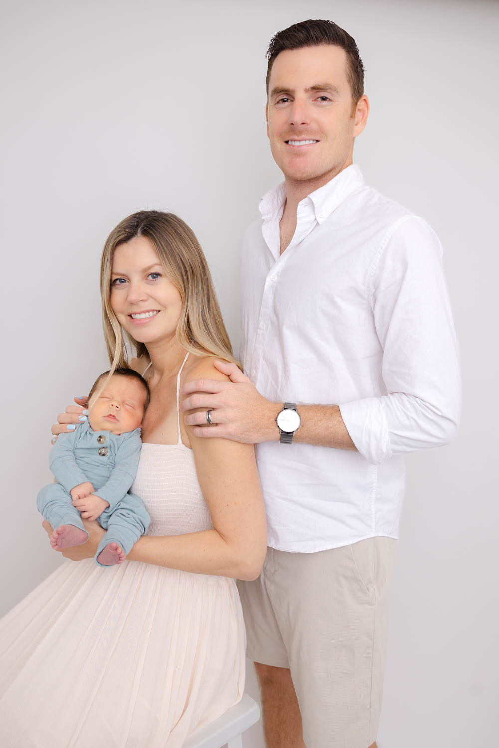 Happy new parents stand and sit in a studio with their newborn against mom's chest in a blue onesie before finding mommy and me classes in redondo beach