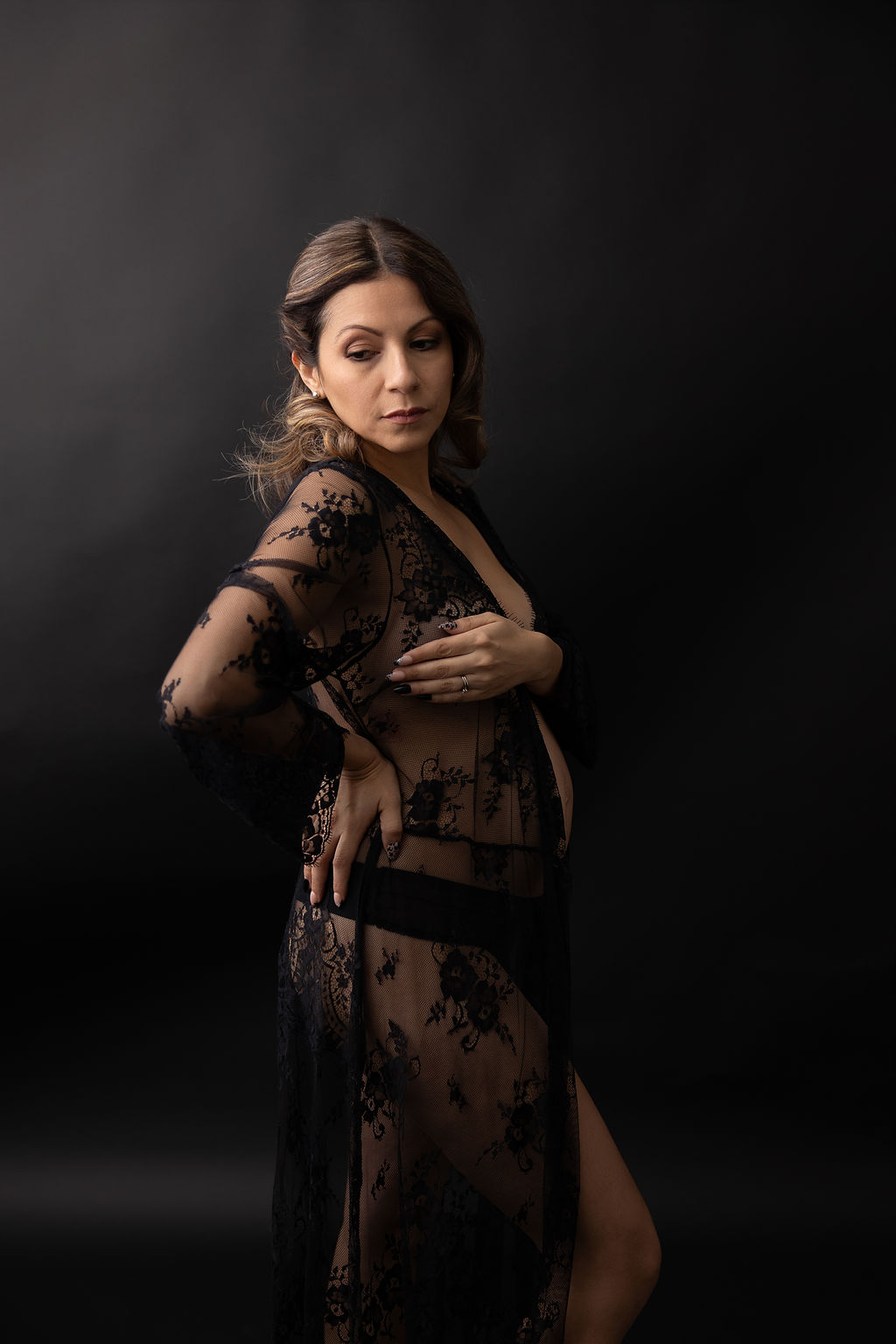 A pregnant woman stands with a hand on her lower back while wearing a sheer black lace robe in a studio