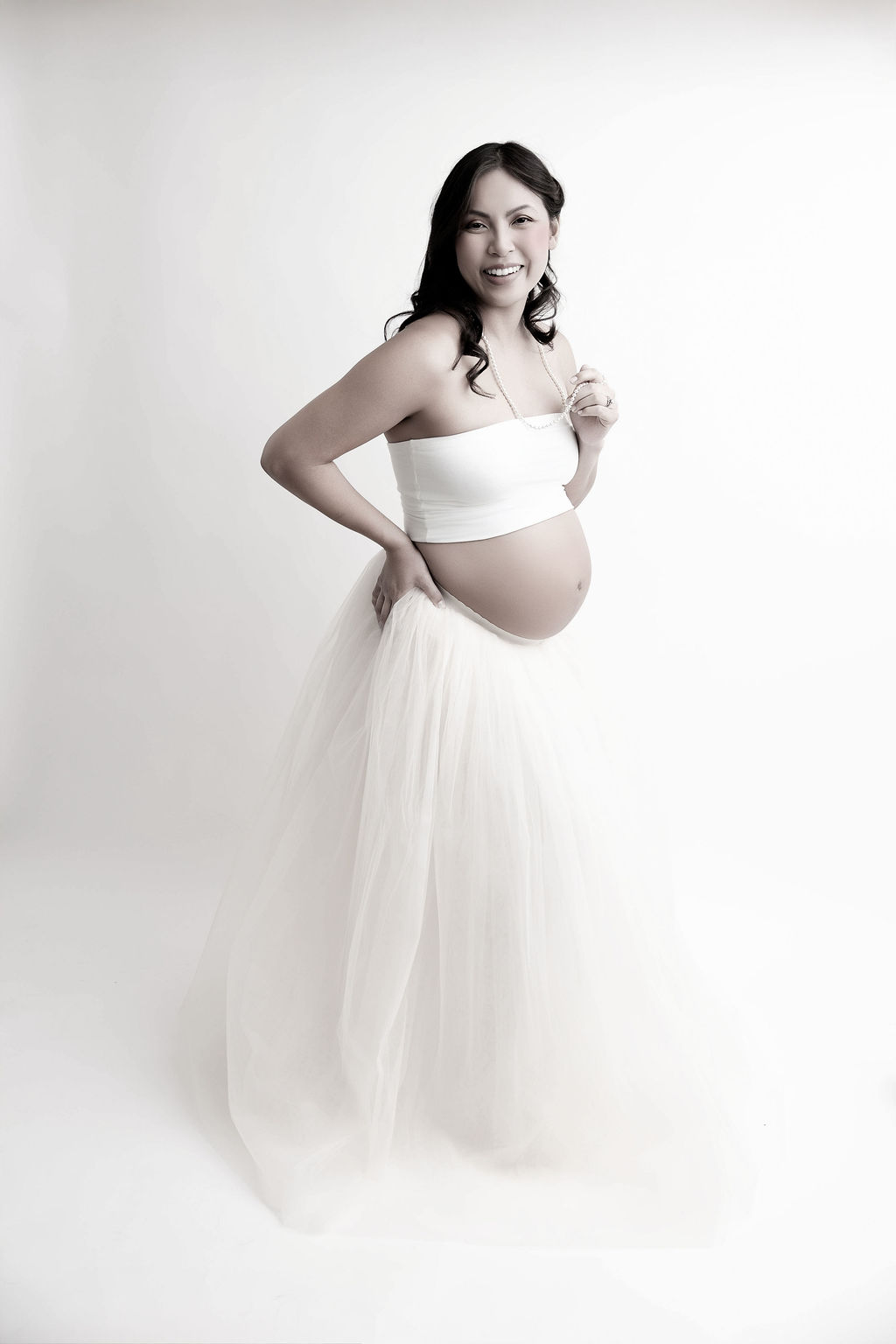A laughing pregnant woman in a tule skirt and white tube top in a studio with a hand on her hip after visiting a prenatal chiropractor in redondo beach