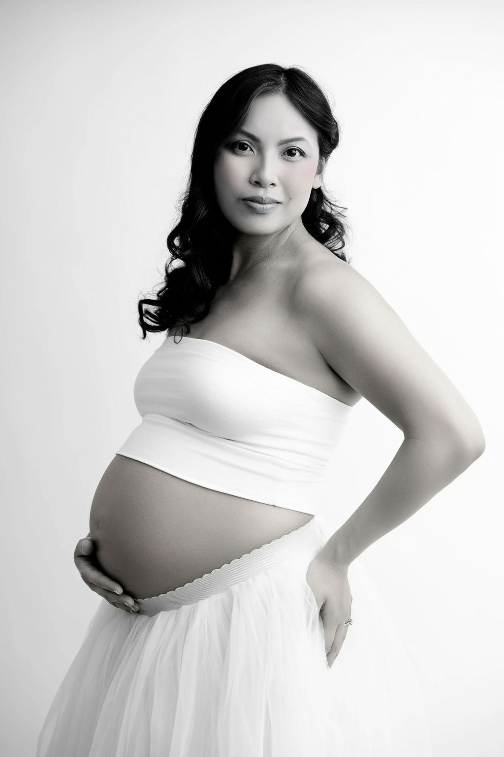 A pregnant woman stands smiling in a studio in a white tube top with a hand under her bump after visiting a prenatal chiropractor in redondo beach