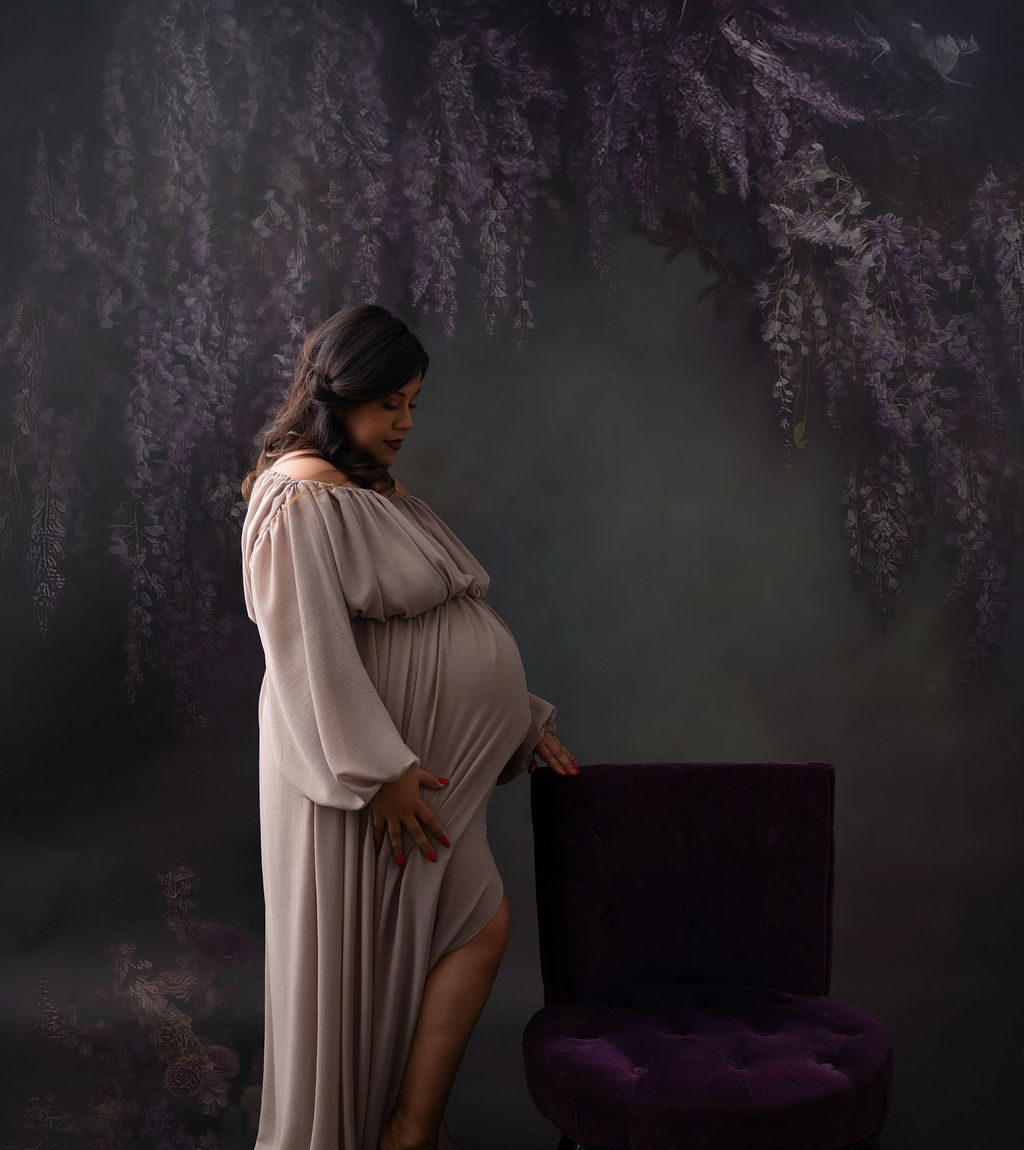 A pregnant woman stands by a purple chair in a studio in a matching maternity gown