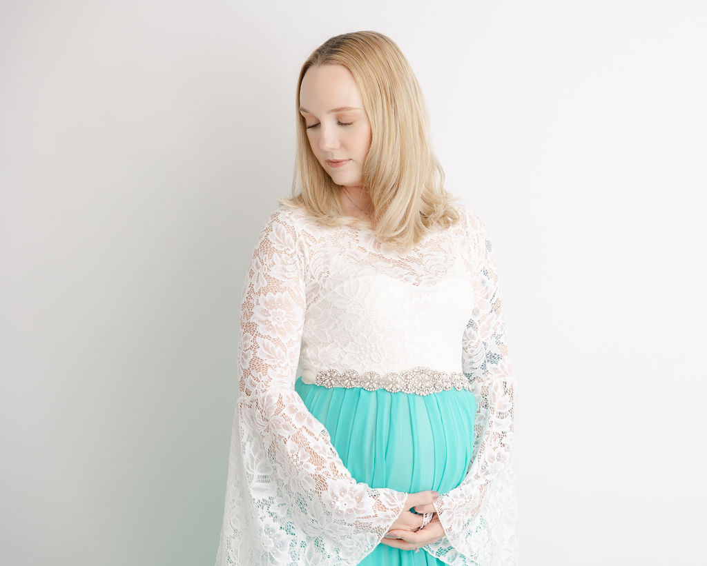 An expecting woman in a lace maternity gown stands in a studio gazing down her shoulder