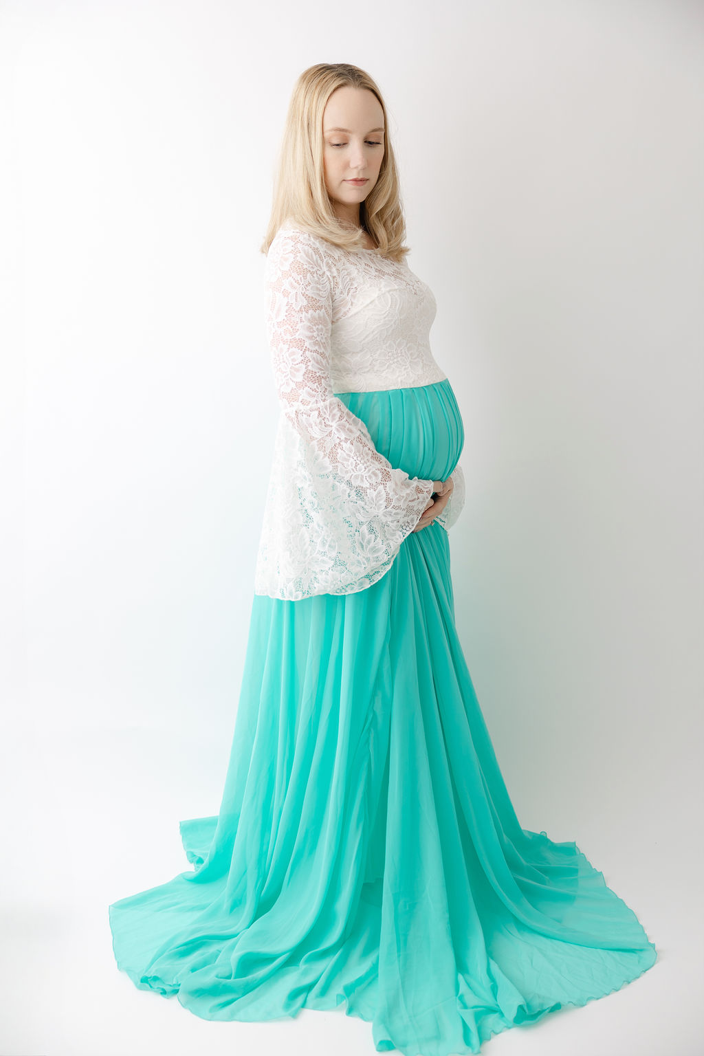 A pregnant woman stands with hands under her bump in a teal maternity gown with lace top after some prenatal yoga in redondo beach
