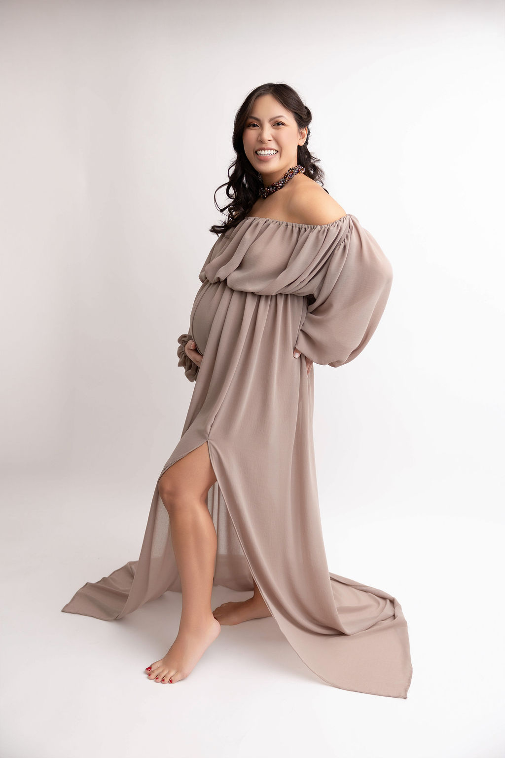 A laughing mom to be in a tan loose maternity gown with a hand under her bump in a studio after her 3d ultrasound in torrance