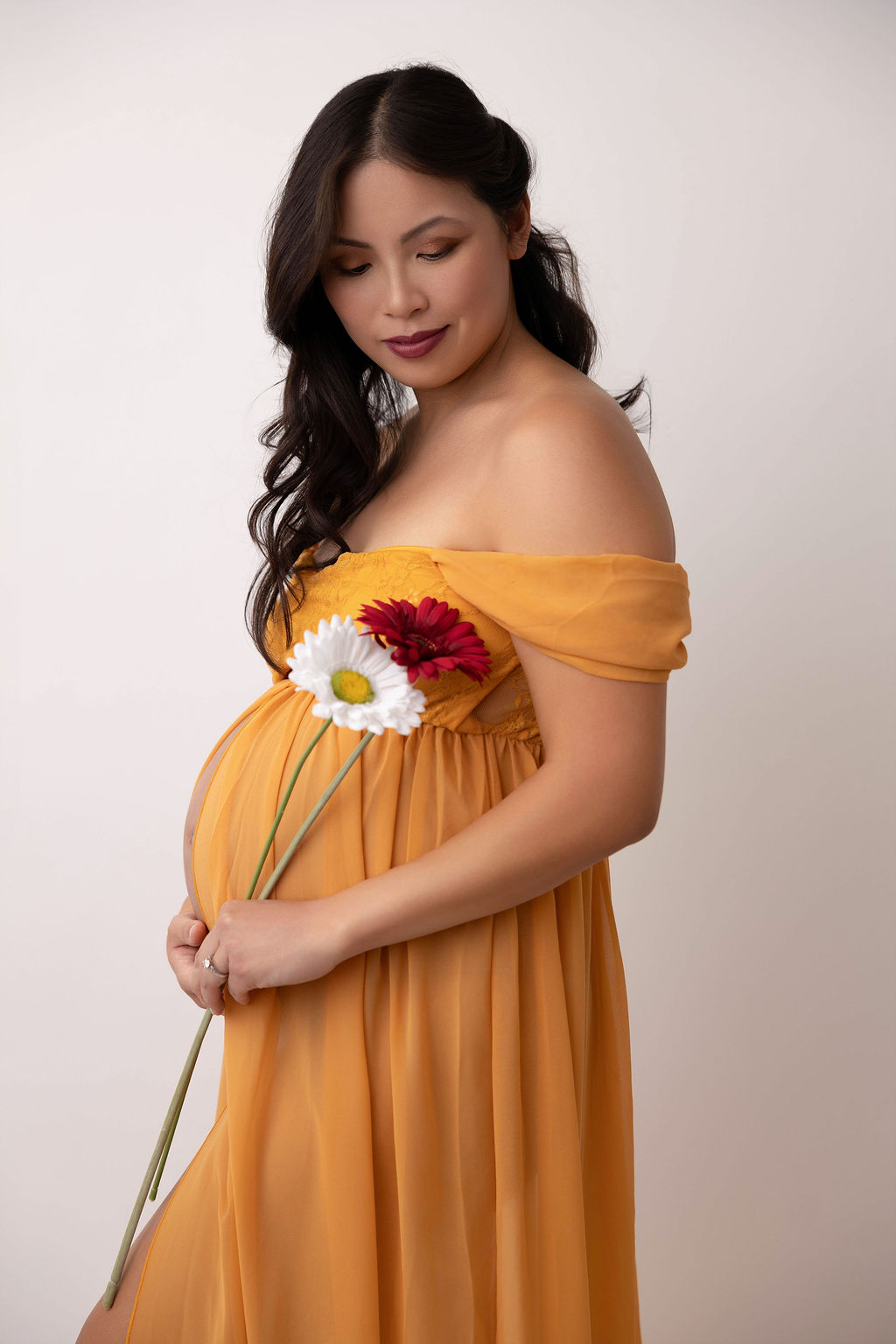 A pregnant woman in a gold maternity gown holds white and red flowers while smiling down her shoulder after visiting birth centers in torrance