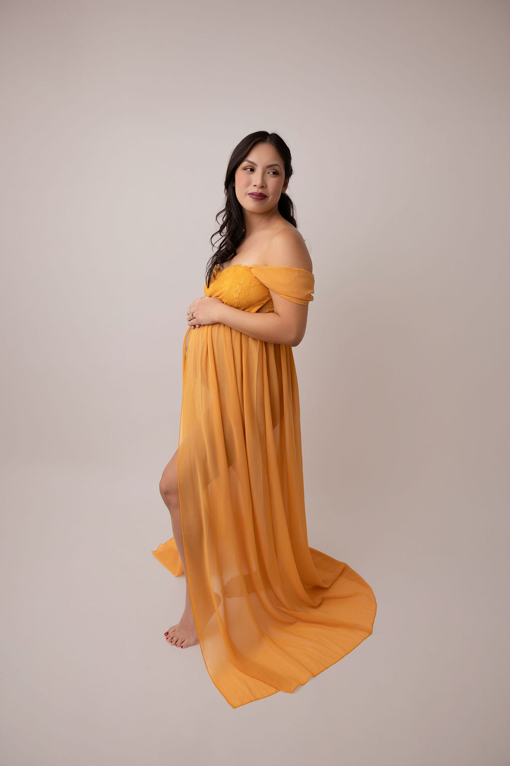 A mother to be in a long gold maternity gown stands smiling over her shoulder in a studio after visiting birth centers in torrance
