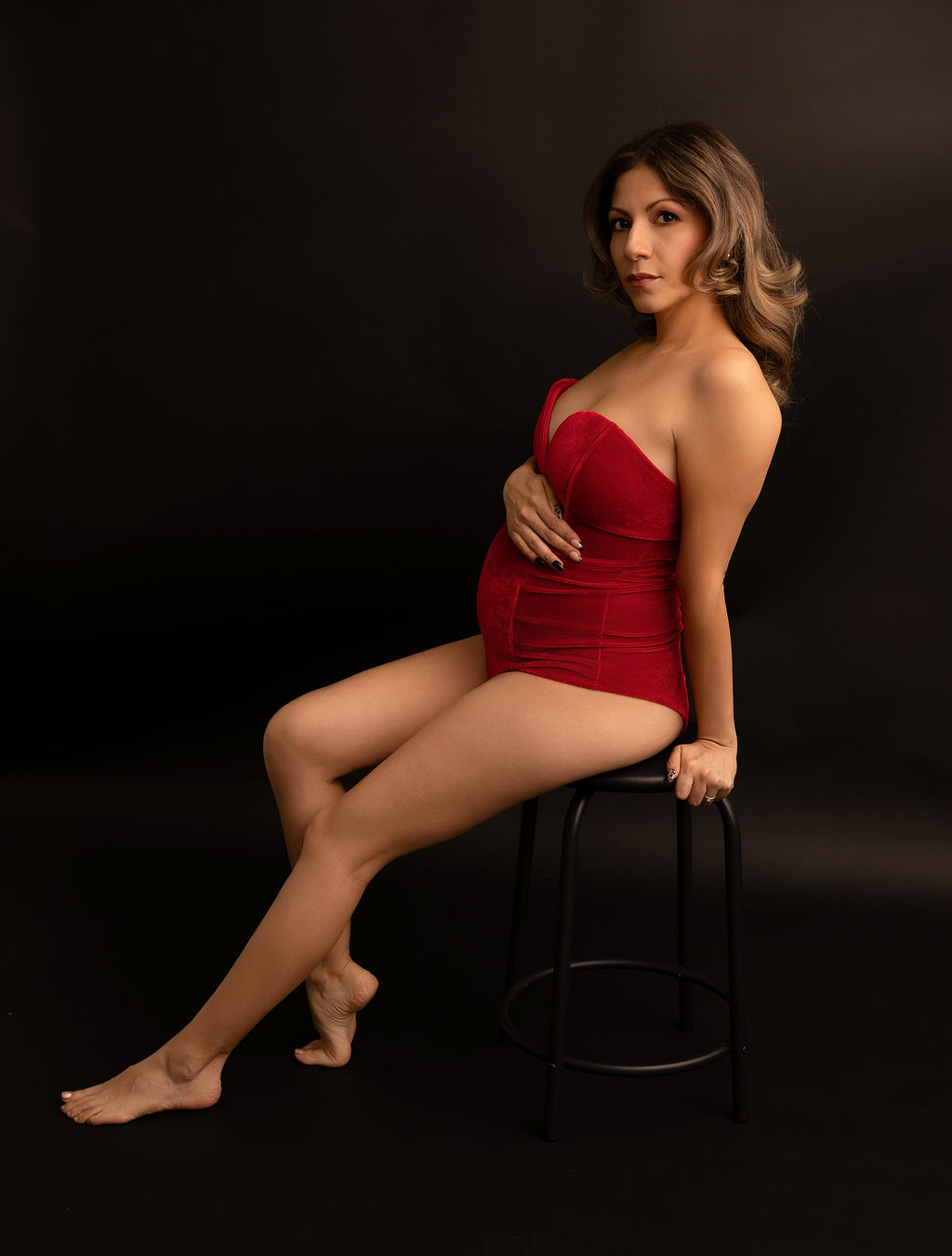 A mother to be in a red maternity bodysuit leans on a stool in a studio with a hand on her bump