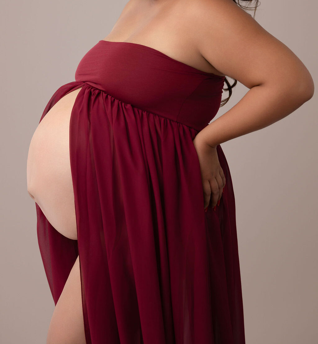 A pregnant woman in an open red maternity dress stands smiling to her bump with hands on her hips after meeting doulas in Torrance