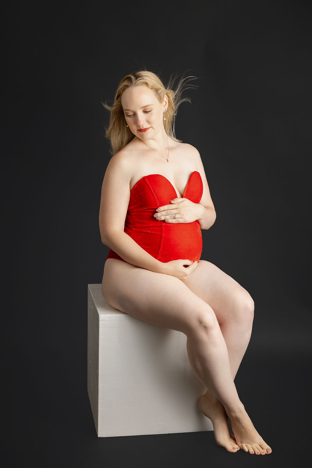 A mother to be in red maternity lingerie sits on a whtie cube ina. studio with hair blowing after meeting doulas in Torrance