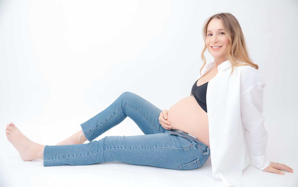 A happy pregnant woman in jeans and white shirt sits across a studio floor smiling