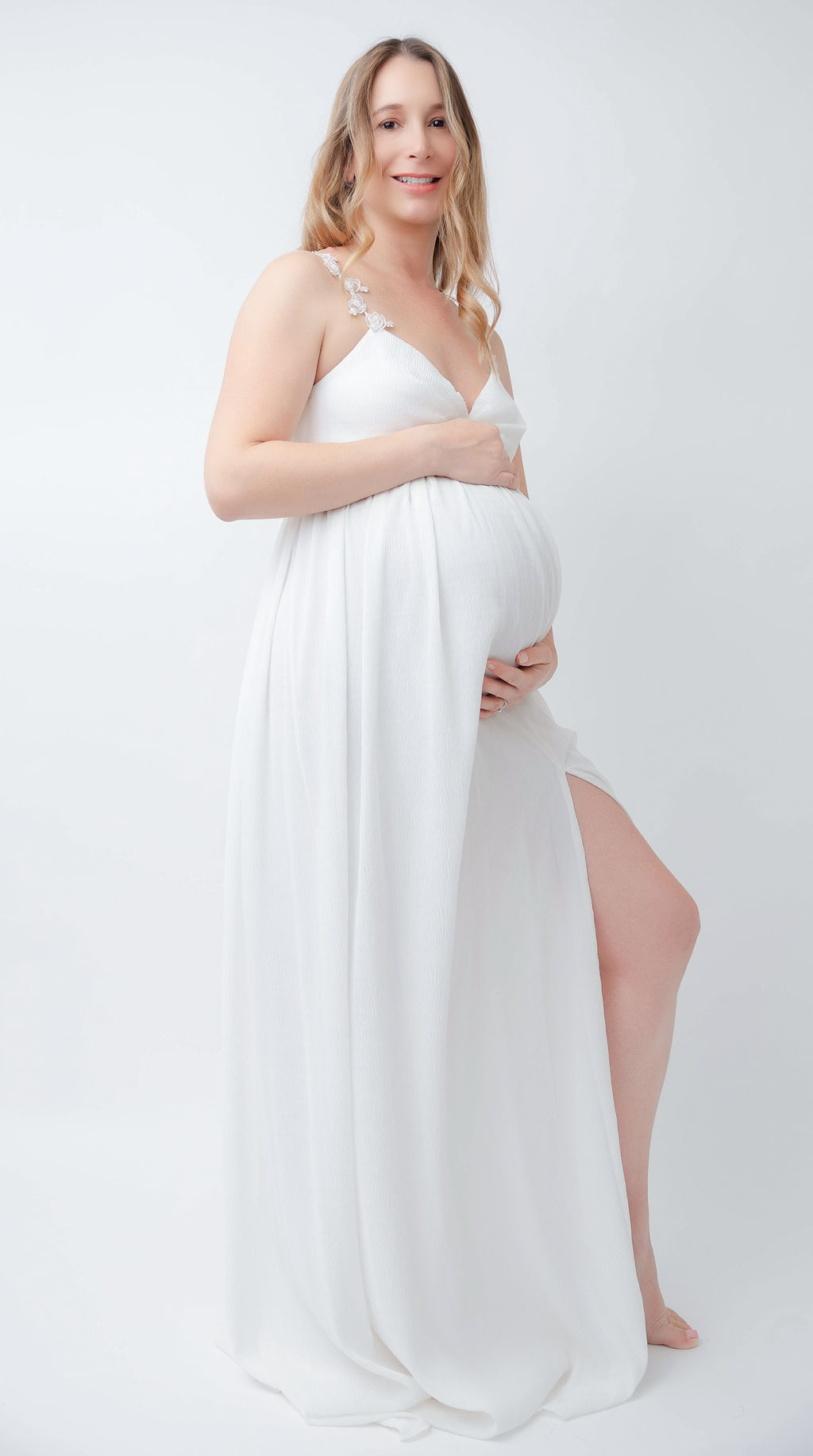A smiling woman in a white maternity gown stands in a studio holding her bump thanks to fertility clinics in torrance