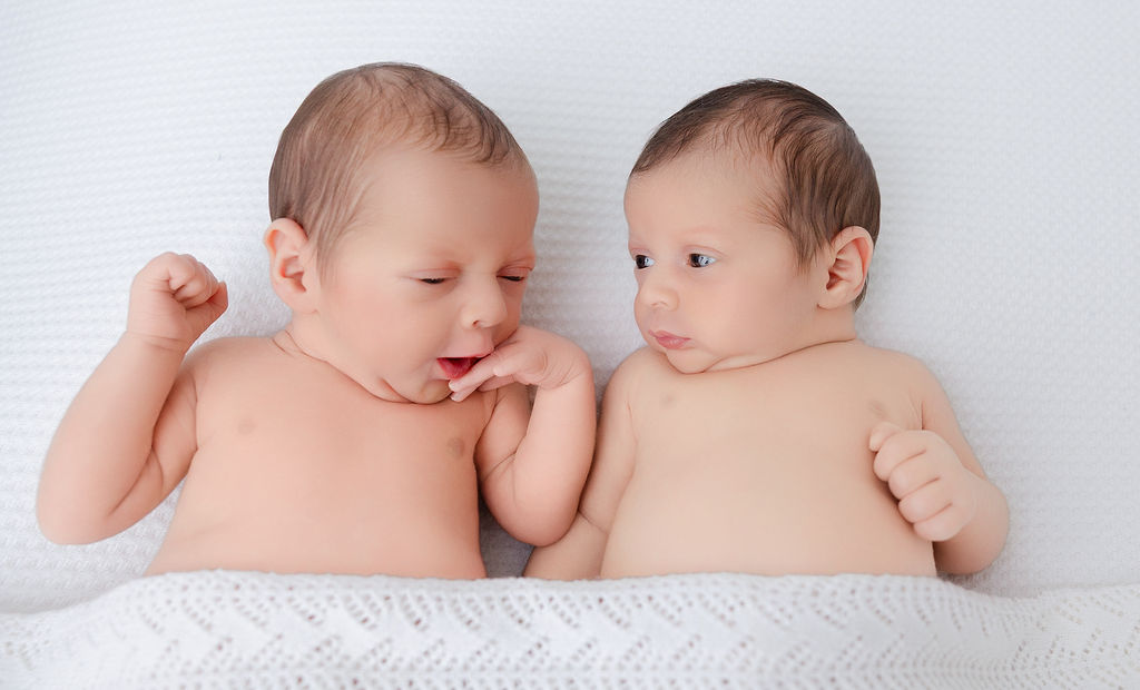 Newborn baby twins lay on a bed under a blanket awake with eyes open