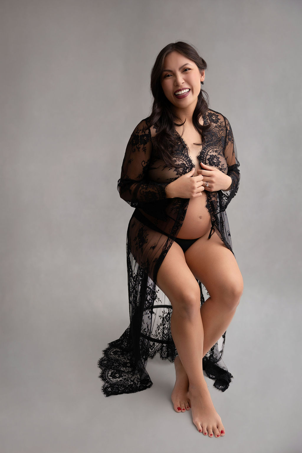 A happy pregnant woman laughs while sitting on a stool in a black lace lingerie coverup after meeting midwives in torrance