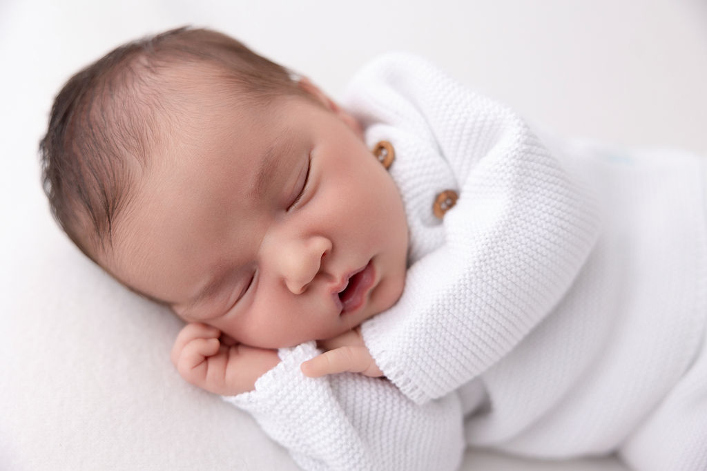 A newborn baby sleeps on its side in a white onesie with hands under cheek