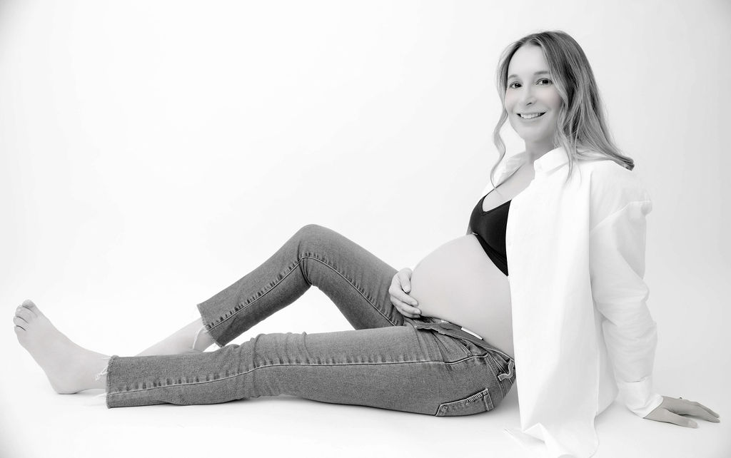 A happy pregnant woman sits on the floor of a studio in open jeans and white shirt after finding obgyns in torrance