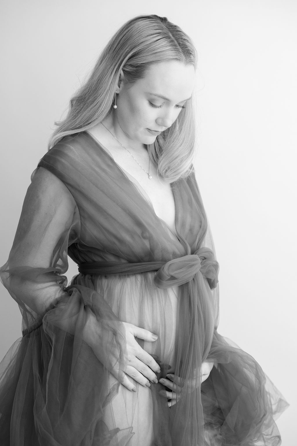 A mother to be stands in a sheer maternity robe looking down to her bump after visiting obgyns in torrance