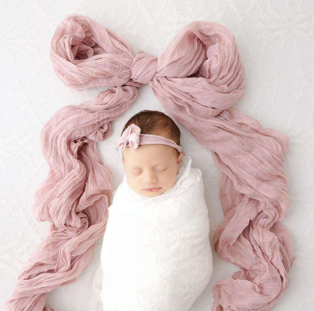 A newborn baby girl sleeps in a white swaddle surrounded by a pink fabric tied in a bow