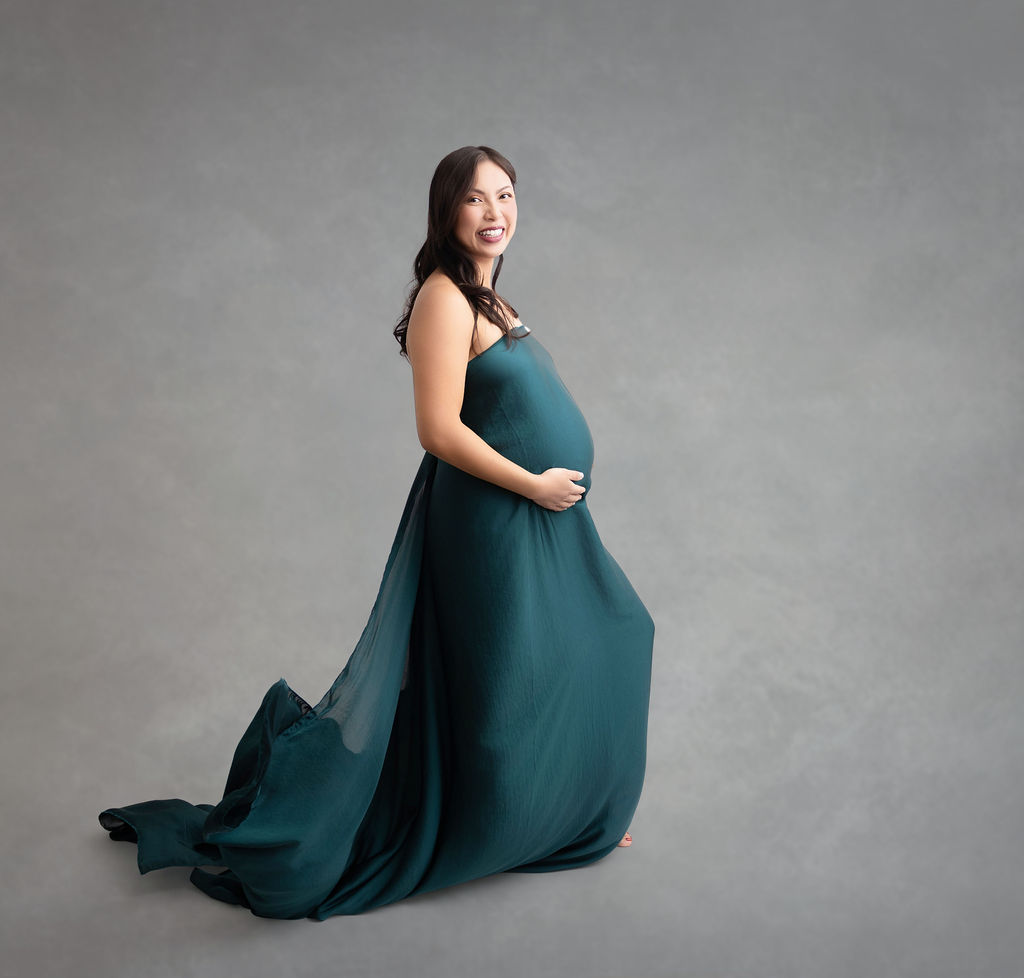A smiling pregnant woman wrapped in a teal silk stands in a studio smiling with hands under her bump