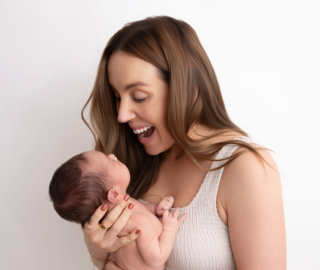 A happy mom laughs while playing with her newborn baby against her chest in a studio