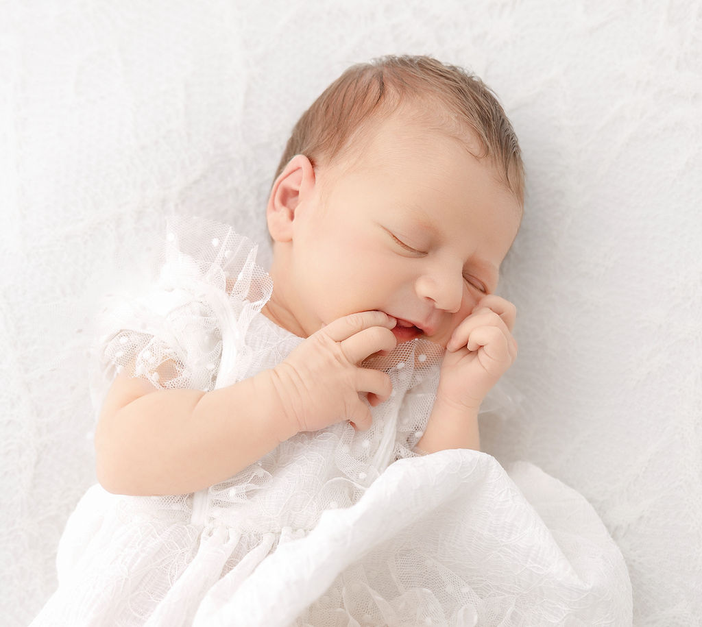 A newborn baby girl in a white gown sleeps with a finger in her mouth on a white bed after meeting pediatric chiropractors in torrance