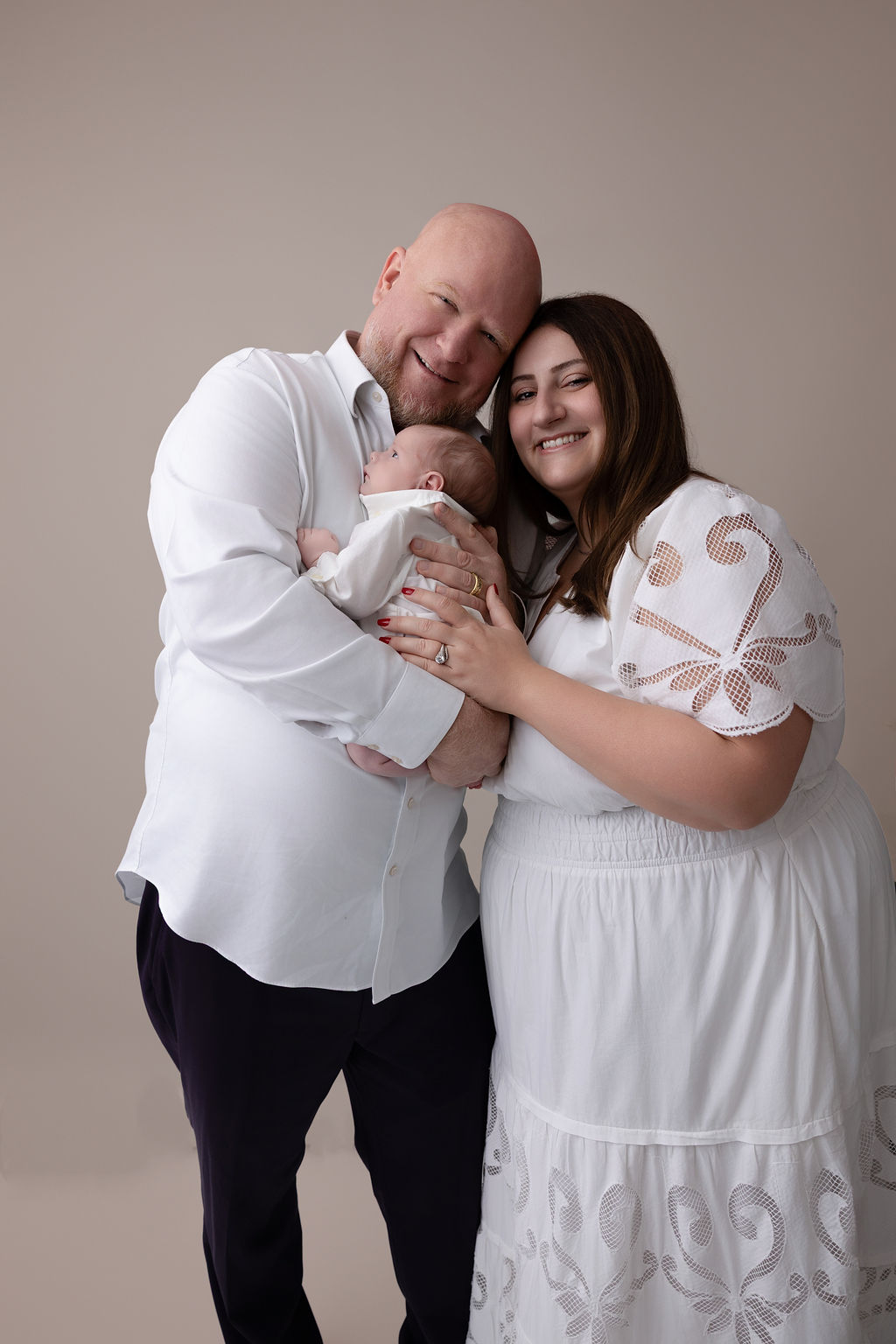 smiling new mom in a white dress snuggles her husband and newborn against his chest while standing in a studio
