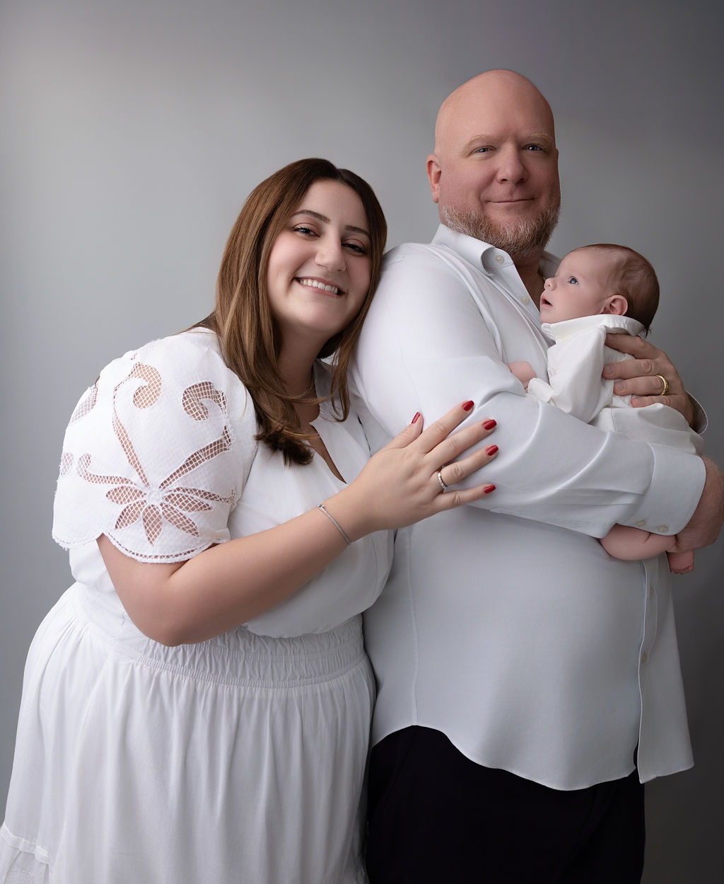 Happy new parents in white stand with their newborn against dad's chest in a studio after some pelvic floor therapy in Torrance
