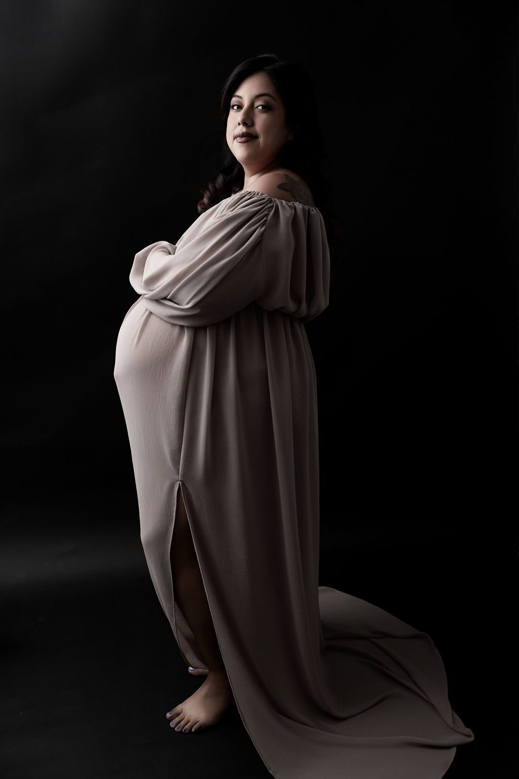 A pregnant woman stands in a beige maternity gown ina. dark studio smiling with hands on her bump