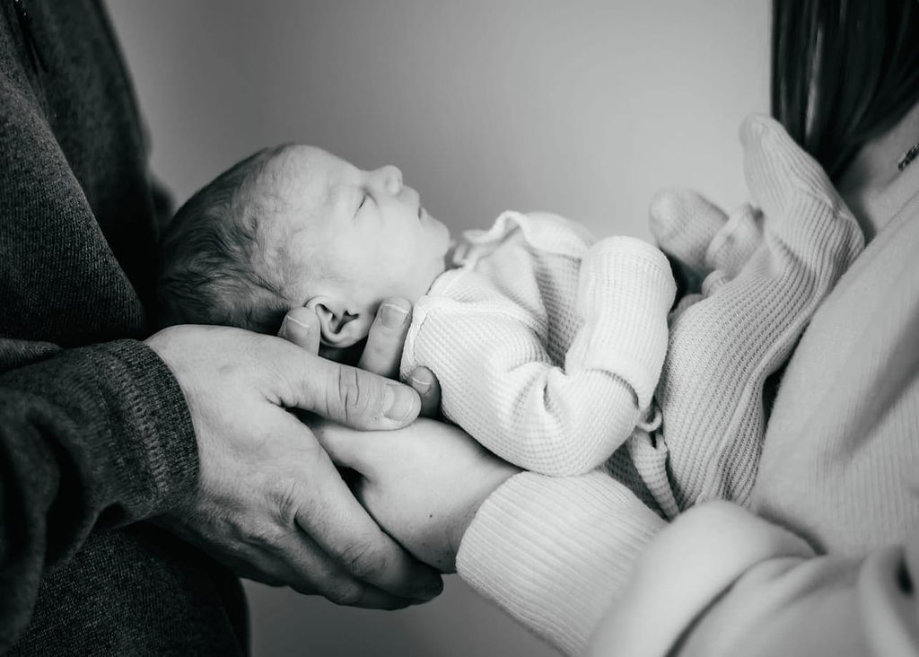 A black and white image of a newborn baby sleeping between mom and dad in their hands in a onesie thanks to prenatal chiropractors in torrance