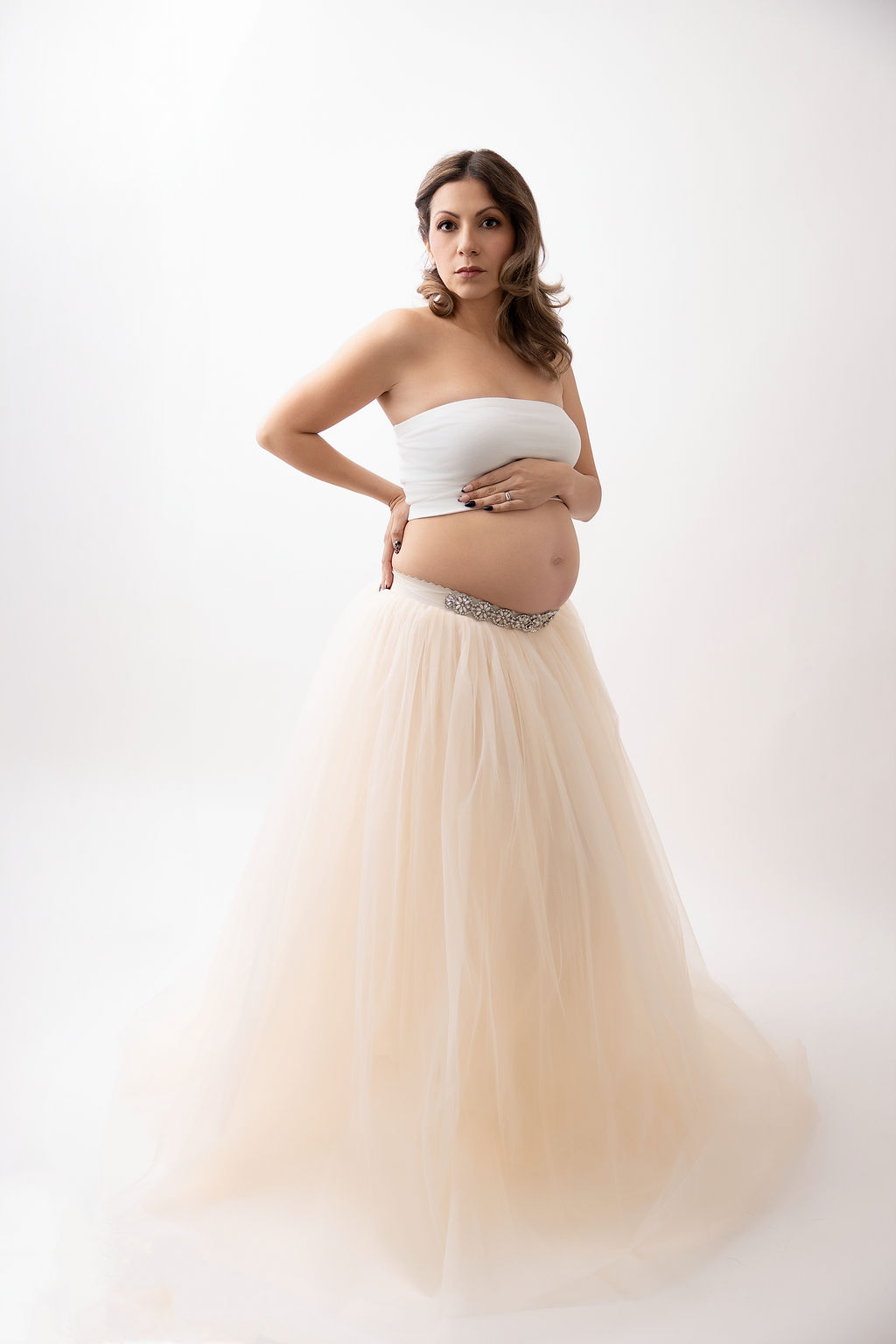 A pregnant woman in a white top and cream skirt stands with a hand on her bump in a studio after a prenatal massage in torrance